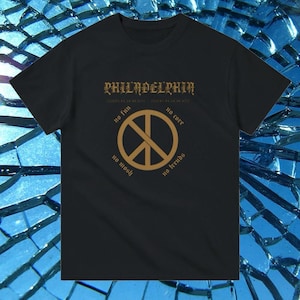 以下が含まれることがあります： 黒いTシャツ。 金色の平和のシンボルとテキストが特徴です。 テキストには、上部に「PHILADELPHIA」と、平和のシンボルの周りに「no fun」、「no core」、「no mosh」、「no trends」などのフレーズが含まれています。