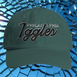 May include: A dark green baseball cap featuring the word "Philadelphia" embroidered in white and "Eagles" in black script. The cap has a curved peak and a classic six-panel design.