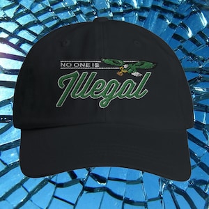 Op de afbeelding: Zwarte baseballcap met de geborduurde tekst "NO ONE IS Illegal" in wit en groen. De cap heeft een groene en witte adelaarsafbeelding. De hoed is gemaakt van een duurzame stof en heeft een gebogen klep.