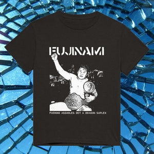 Puede incluir: Camiseta negra con un gráfico blanco de un luchador sosteniendo un cinturón de campeonato. El texto "FUJINAMI" está encima del luchador, y la frase "FUCKING ASSHOLES GET A DRAGON SUPLEX" está debajo.