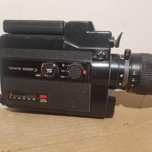 Może przedstawiać: Czarna kamera filmowa Super 8 Canon 514XL z obiektywem Canon Zoom Lens C-8. Panel boczny zawiera wskaźnik naładowania baterii, pokrętło samowyzwalacza oraz przełączniki. Korpus aparatu posiada teksturowany uchwyt oraz wyraźny tubus obiektywu z pierścieniami.