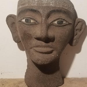 May include: A weathered, brown stone sculpture of a human head. The face has detailed features, including eyes with painted irises, a defined nose, and a subtle smile. The head is adorned with a band across the forehead, suggesting ancient artistry.