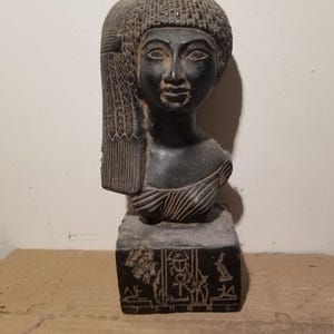 May include: A dark-coloured bust of an Egyptian figure, featuring a detailed headdress and facial features. The bust is mounted on a square base with hieroglyphic carvings. The sculpture is made of a dark, possibly stone material.