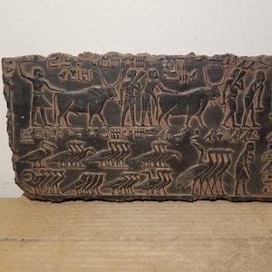 May include: A dark, rectangular stone carving featuring ancient Egyptian hieroglyphs and figures. The relief depicts people, cattle, and birds, with detailed carvings and a weathered appearance. The artwork is a historical artifact.