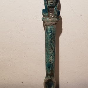 May include: A teal-coloured ceramic cosmetic spoon featuring a carved human head at the top, reminiscent of ancient Egyptian style. The elongated handle is decorated with vertical hieroglyphic-style engravings, and the base forms a small, shallow bowl.