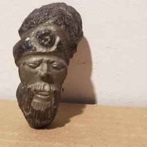 Puede incluir: Una escultura metálica de tonos oscuros que representa una cabeza con rostro barbudo y ojos cerrados. La figura lleva un tocado redondeado y texturizado adornado con un emblema circular central. El objeto tiene un aspecto envejecido y patinado.