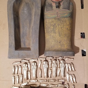 May include: A large, hollow, dark grey stone sarcophagus base next to a matching lid painted with a human face, a colourful collar, and yellow hieroglyphics. Below are numerous small, metallic gold-coloured miniature sarcophagus figurines.