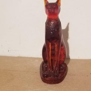 Puede incluir: Una figura de cristal translúcido de color ámbar intenso que representa a un gato sentado. El gato aparece en una postura erguida y alerta, con orejas puntiagudas y un diseño de collar detallado alrededor del cuello. El cristal es liso.