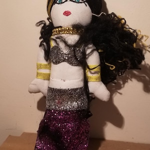 May include: Handmade doll with a white face, blue eyes, and red lips. The doll has black and yellow yarn hair, a gold beaded necklace, and a glittery silver top and skirt. Gold armbands complete the outfit.