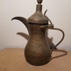 May include: A tarnished brass coffee pot featuring a bulbous body, a long curved spout, and a hinged lid topped with a finial. The vessel includes a slender handle attached to the side, showcasing a weathered metallic finish with visible patina.