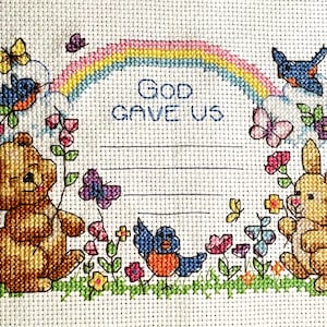 May include: Cross-stitch design featuring a rainbow, butterflies, birds, a teddy bear, and a bunny. The text "GOD GAVE US" is stitched above a blank space. The design is surrounded by flowers and greenery.