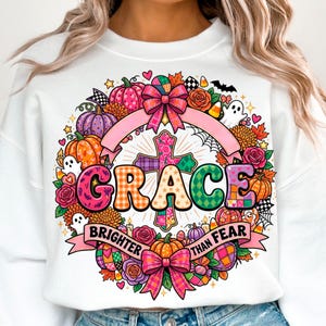 May include: A white crewneck sweatshirt featuring a colorful graphic print of a cross surrounded by pumpkins, flowers, ghosts, and bows. The text reads GRACE in large letters and BRIGHTER THAN FEAR on a pink ribbon banner below the design.
