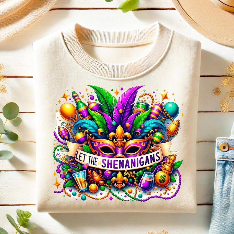May include: Cream-colored sweatshirt featuring a vibrant Mardi Gras design. The graphic includes a mask, feathers, beads, and the text "LET THE SHENANIGANS begin". Colorful bottles, drinks, and stars complete the festive look.