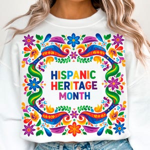 May include: White sweatshirt with a vibrant floral design in a square frame. The design includes colorful flowers, birds, and the text "HISPANIC HERITAGE MONTH" in a rainbow of colors. The sweatshirt is a casual garment.