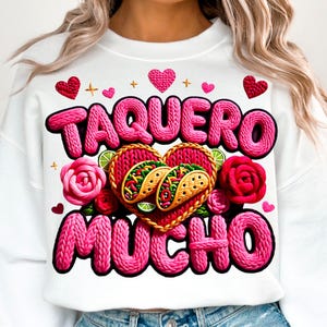 May include: White sweatshirt with the text "TAQUERO MUCHO" in pink, featuring a heart-shaped design with tacos, roses, and hearts. The design is in a playful, cartoon style, perfect for a casual look.