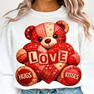Può includere: Felpa bianca con un orsacchiotto patchwork che tiene un cuore. Il cuore dice "LOVE" e l'orso ha "HUGS" e "KISSES" sulle zampe. L'orso è rosso, beige e bianco, con un design cucito.