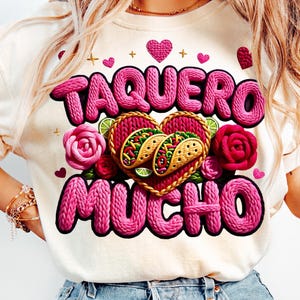 Puede incluir: Camiseta color crema con las palabras "TAQUERO MUCHO" en letras rosas texturizadas. El dise&ntilde;o incluye corazones rosas, rosas y dos tacos dentro de un dise&ntilde;o tejido en forma de coraz&oacute;n. Tambi&eacute;n se incluyen rodajas de lima.