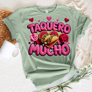 Puede incluir: Camiseta verde claro con la frase "TAQUERO MUCHO" en rosa, con letras texturizadas. El dise&ntilde;o incluye un gr&aacute;fico de tacos en forma de coraz&oacute;n, rosas rosas y peque&ntilde;os corazones. La camiseta est&aacute; anudada en la cintura.