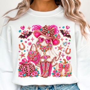 May include: A white sweatshirt featuring a graphic of a ghost wearing a pink leopard print cowboy hat, heart-shaped sunglasses, a jeweled necklace, a fringed handbag, and matching boots, surrounded by pumpkins, bats, and sparkling hearts.