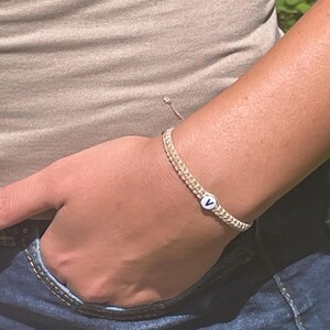 May include: A woven bracelet with a small white bead featuring the letter "V" in black. The bracelet is beige and is worn on a wrist. The person is wearing blue jeans and a beige shirt.