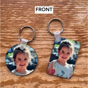 May include: Two keychains with round and rectangular photo inserts. Both keychains feature a photo of a young girl with brown hair smiling. The round keychain is silver and the rectangular keychain is silver with a black border.