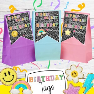 May include: Three colorful party favor bags with black glitter and gold lettering that reads "Hip Hip Hooray It's your Birthday". Each bag has a different design: a smiley face, a rainbow, and a star. The bags are on a white wood background with a sign that reads "Birthday Tags".