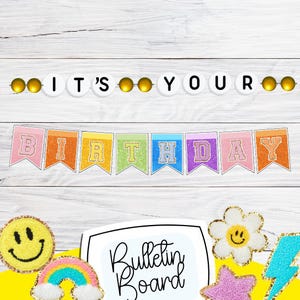 May include: A colorful birthday banner with the words "It's Your Birthday" and a string of white lights with gold bulbs. The banner is made of rainbow-colored felt with glitter letters. The background is a white wooden surface.