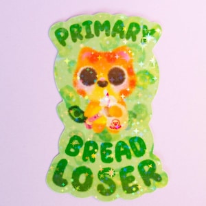 May include: A sticker with a cartoon cat holding a drink, with the words "PRIMARY" and "BREAD LOSER" in green. The cat is orange and yellow with large eyes, set against a green background with glitter.