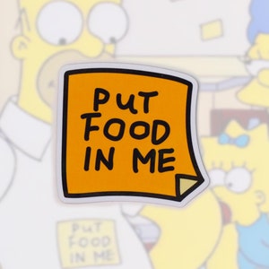 May include: An orange sticker with the text "PUT FOOD IN ME" in black, with a black border. The sticker is in front of a blurred background featuring cartoon characters from The Simpsons.