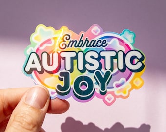 Pode incluir: Um autocolante colorido com as palavras "Embrace AUTISTIC JOY" numa fonte divertida. O autocolante tem uma borda colorida do arco-íris e é segurado contra um fundo roxo.