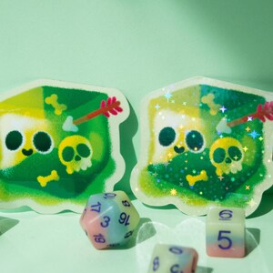 May include: Two green and yellow holographic stickers with a cartoon character design. The character is a green and yellow creature with a skull and crossbones on its chest. The stickers are surrounded by dice with different numbers on them.