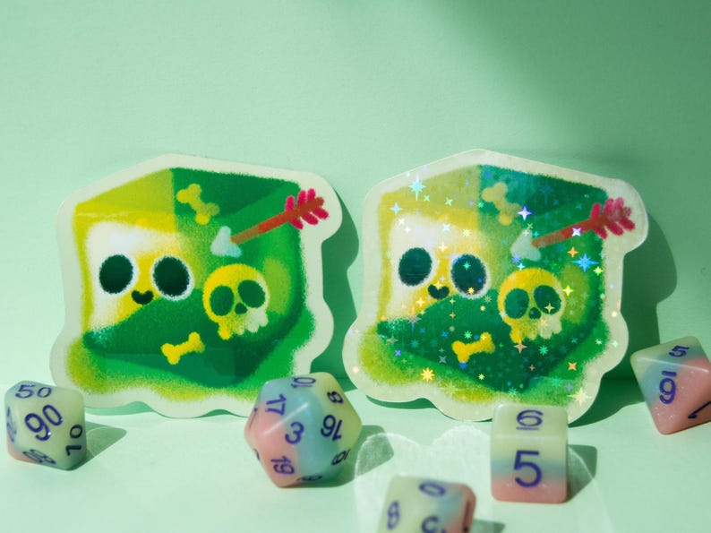 May include: Two green and yellow holographic stickers with a cartoon character design. The character is a green and yellow creature with a skull and crossbones on its chest. The stickers are surrounded by dice with different numbers on them.