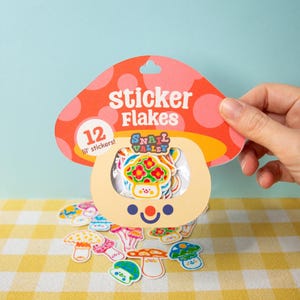 Może przedstawiać: Opakowanie "Sticker Flakes" z 12 małymi naklejkami, trzymane w dłoni. Opakowanie ma kształt grzyba z czerwono-różowym wzorem w kropki. Naklejki mają motyw grzybów w różnych kolorach i wzorach. Kilka naklejek jest rozrzuconych na żółtej kratowanej powierzchni.