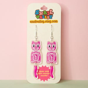Peut inclure: Une paire de boucles d'oreilles pendantes en forme de chat rose en acrylique, avec un nœud papillon blanc et une queue séparée. Les boucles sont fixées sur une carte de présentation avec le texte Snail Valley et snailvalley.etsy.com.