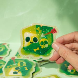 May include: A green and yellow glitter sticker with a cartoon skull and crossbones design. The sticker is holographic and has a sparkly finish.