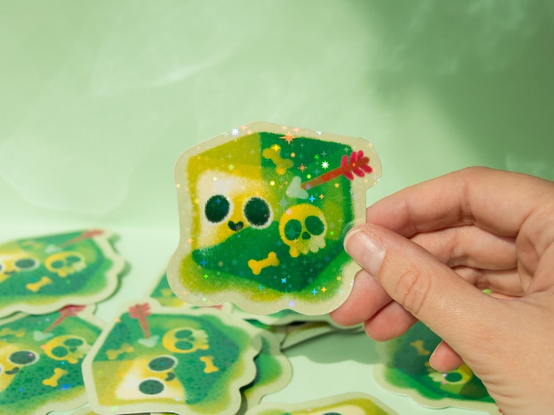 May include: A green and yellow glitter sticker with a cartoon skull and crossbones design. The sticker is holographic and has a sparkly finish.
