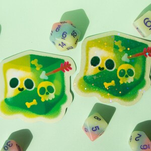 May include: Two green and white dice stickers with glitter and a cartoon skull and arrow design. The stickers are on a light green background with other dice.