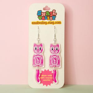 May include: Pink cat-shaped earrings with silver hooks, displayed on a cream card. The earrings feature a gummy bear design with white details. The card has the text "Snail Valley etsy.com" and "Made with 100% recycled acrylic!"