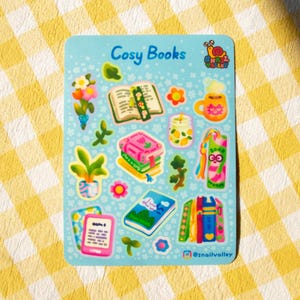 May include: A sticker sheet with a light blue background featuring illustrations of books, flowers, and a mug. The text "Cosy Books" is at the top. The @snailvalley logo is at the bottom. The sheet is decorated with small white dots.