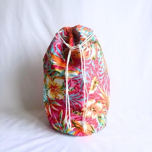 May include: A drawstring bag featuring a bright floral design in pink, orange, yellow, and blue. The bag is made of fabric and has a white drawstring closure. The overall design is tropical and colourful.