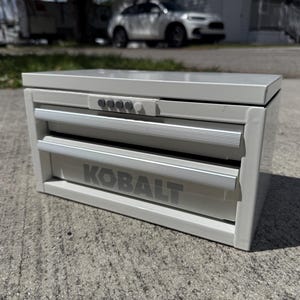 May include: A light gray metal tool chest with two drawers and a top compartment. The word "KOBALT" is printed in large, gray letters on the bottom drawer. The drawers have silver-colored handles.