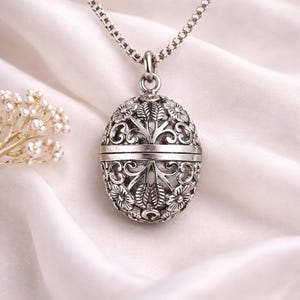 Può includere: Collana con pendente color argento con un medaglione ovale con intricati motivi floreali e a foglie. Il medaglione pende da una delicata catena. Lo sfondo è un morbido tessuto bianco.