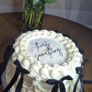 Puede incluir: Un pastel glaseado blanco con las palabras "thirty something" escritas en la parte superior. El pastel está decorado con lazos y cintas negras. Un jarrón de cristal con tallos y hojas verdes está en el fondo.