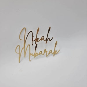 May include: Gold metallic calligraphy script that reads "Nikah Mubarak" on a white background.