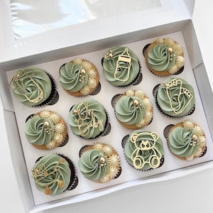 Può includere: Una scatola di 12 cupcake verdi e dorati decorati con topper a tema baby. I topper includono un biberon, una tutina, un orsacchiotto, un'impronta di piede e le parole "Oh Baby".