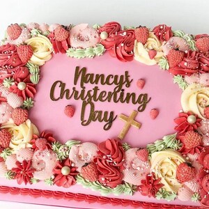 May include: A square pink cake decorated with pink, white and green frosting. The cake is decorated with a border of flowers and strawberries. The cake has a gold inscription that reads "Nancy's Christening Day" and a gold cross.