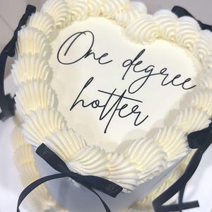 Può includere: Una torta bianca a forma di cuore con fiocchi di nastro nero e glassa bianca. La torta è decorata con un bordo a tubo e le parole "One degree hotter" sono scritte in glassa nera.