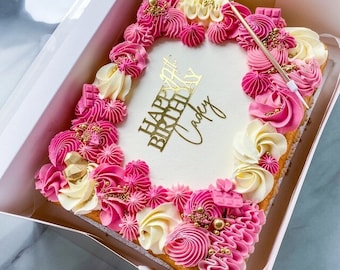 Op de afbeelding: Vierkante taart versierd met roze en witte frosting, gouden sprinkles en een gouden topper met de tekst "Happy Birthday Cadey".