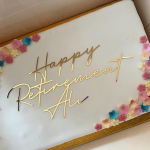 Könnte beinhalten: Ein rechteckiger weißer Kuchen mit goldenem Schriftzug "Happy Retirement Ali". Der Kuchen ist mit kleinen, bunten Blumendekorationen in Rosa, Blau und Gelb an den Rändern verziert.