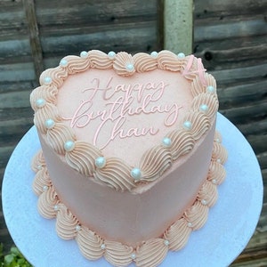 Op de afbeelding: Een roze hartvormige taart versierd met witte parelsprinkels en de woorden "Happy Birthday Chan" in roze glazuur.